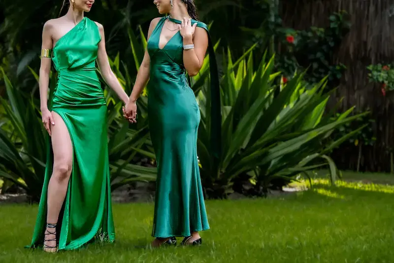 Two elegant Bucharest high class escorts in green satin evening gowns holding hands in a lush garden