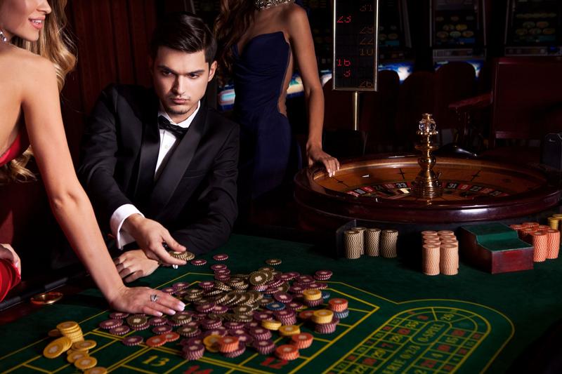 Man in a tuxedo placing a bet at a roulette table with two companions in evening dresses