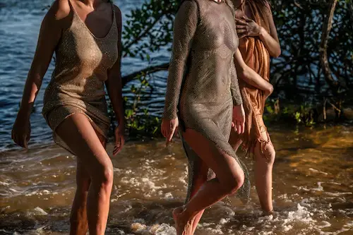 Three elegant women in shimmering evening dresses walking along the water