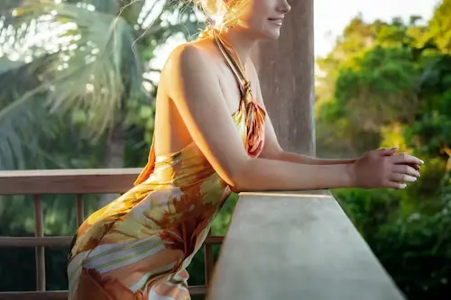 Blonde woman in a flowing orange and gold dress relaxing on a sunlit tropical villa terrace